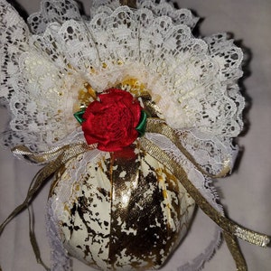 May include: A gold and white fabric Christmas ornament with a red rose and white lace trim.