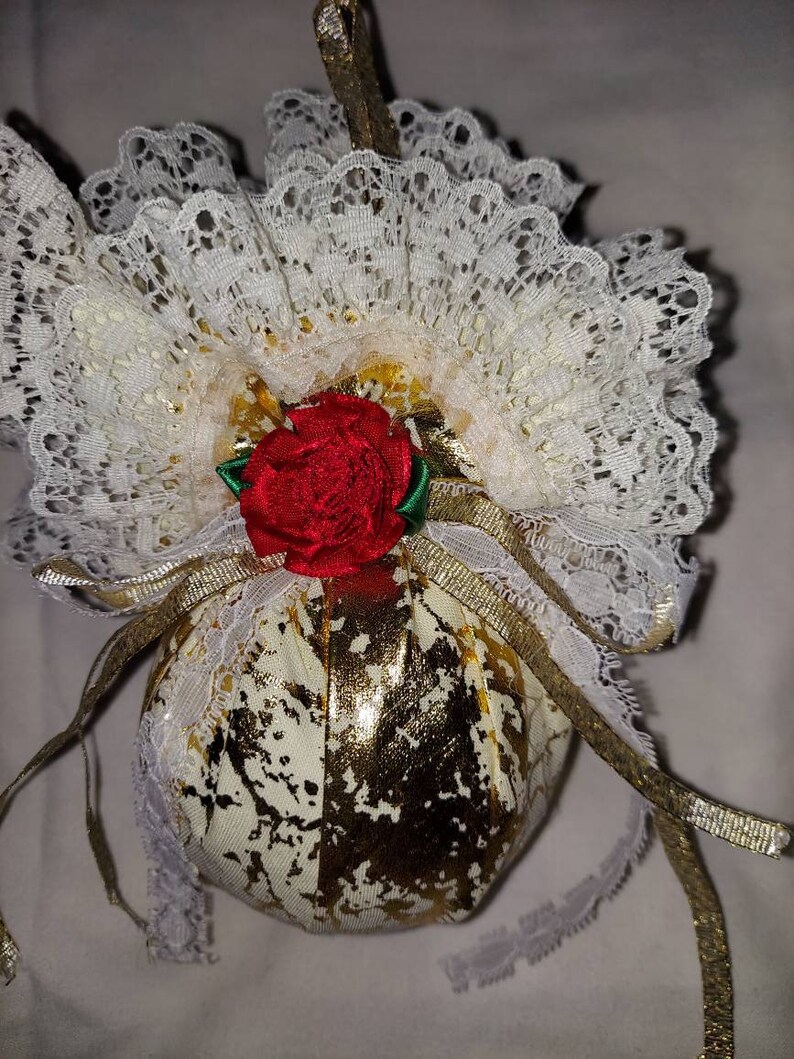 May include: A gold and white fabric Christmas ornament with a red rose and white lace trim.