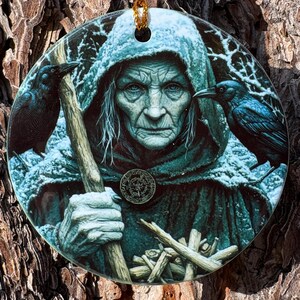 May include: Round ceramic ornament featuring a detailed illustration of an elderly woman in a hooded cloak, holding a staff and surrounded by ravens. The ornament has a gold string for hanging and is set against a textured tree bark background.