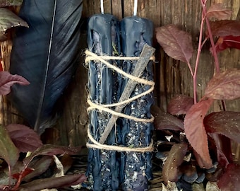 May include: Two dark blue pillar candles with a textured, dripped wax finish, bound together by thin tan twine. A single metal nail is tucked diagonally under the twine across the front of the candles. Each candle features a white wick at the top.
