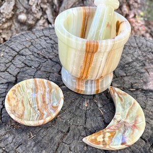 May include: A green, brown, and white onyx mortar and pestle set with a small round dish and a crescent moon-shaped dish. The mortar has a rounded base and a matching pestle. The items are displayed on a weathered wooden surface.