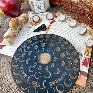 May include: A dark blue wooden planchette with engraved moon phases, numbers, and the words "YES", "NO", and "MAYBE". The image also includes a red apple, a jar of bath soak, candles, and autumn-themed decorations.