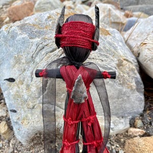 May include: A handcrafted doll with a black head and body, adorned with red wrappings and a red blindfold. The doll features a stone arrowhead and black sheer fabric accents, set against a natural, rocky backdrop.
