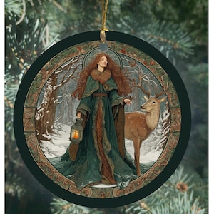 May include: Round Christmas ornament featuring a woman in a green cloak holding a lantern, standing with a deer in a snowy forest scene. The ornament has a gold string for hanging and a dark green border with decorative elements.