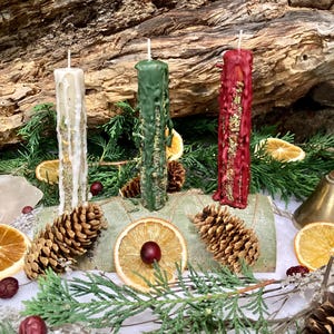 May include: Three pillar candles in white, green, and red, each featuring wax drips and embedded dried herbs. The candles are mounted on a cylindrical wooden base decorated with pinecones, dried orange slices, and red berries.