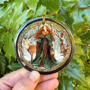 May include: Round Christmas ornament featuring a woman with red hair and a green dress, holding a lantern, standing next to a deer in a snowy forest scene. The ornament has a gold hanging loop and a black border.