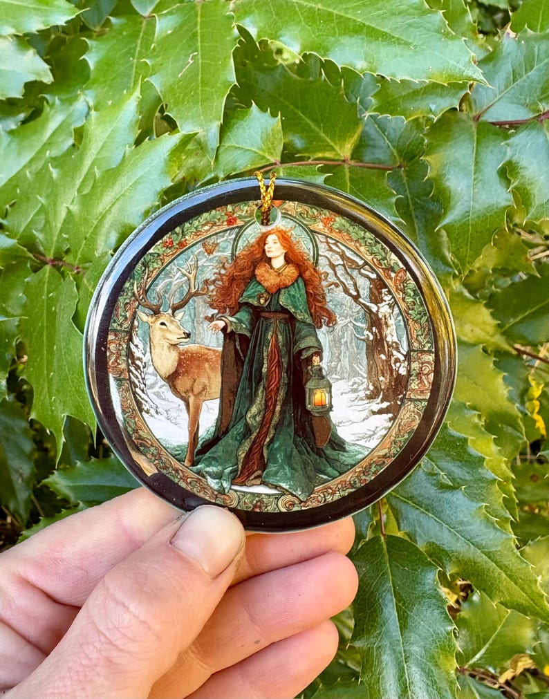 May include: Round Christmas ornament featuring a woman with red hair and a green dress, holding a lantern, standing next to a deer in a snowy forest scene. The ornament has a gold hanging loop and a black border.