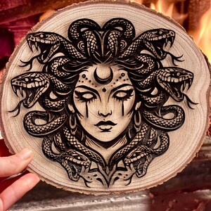 Op de afbeelding: Een houtgebrand kunstwerk met het gezicht van Medusa, met slangen als haar. Het kunstwerk is uitgesneden in een ronde, lichtgekleurde houten schijf. De slangen zijn gedetailleerd en het gezicht heeft een halve maan boven het voorhoofd.