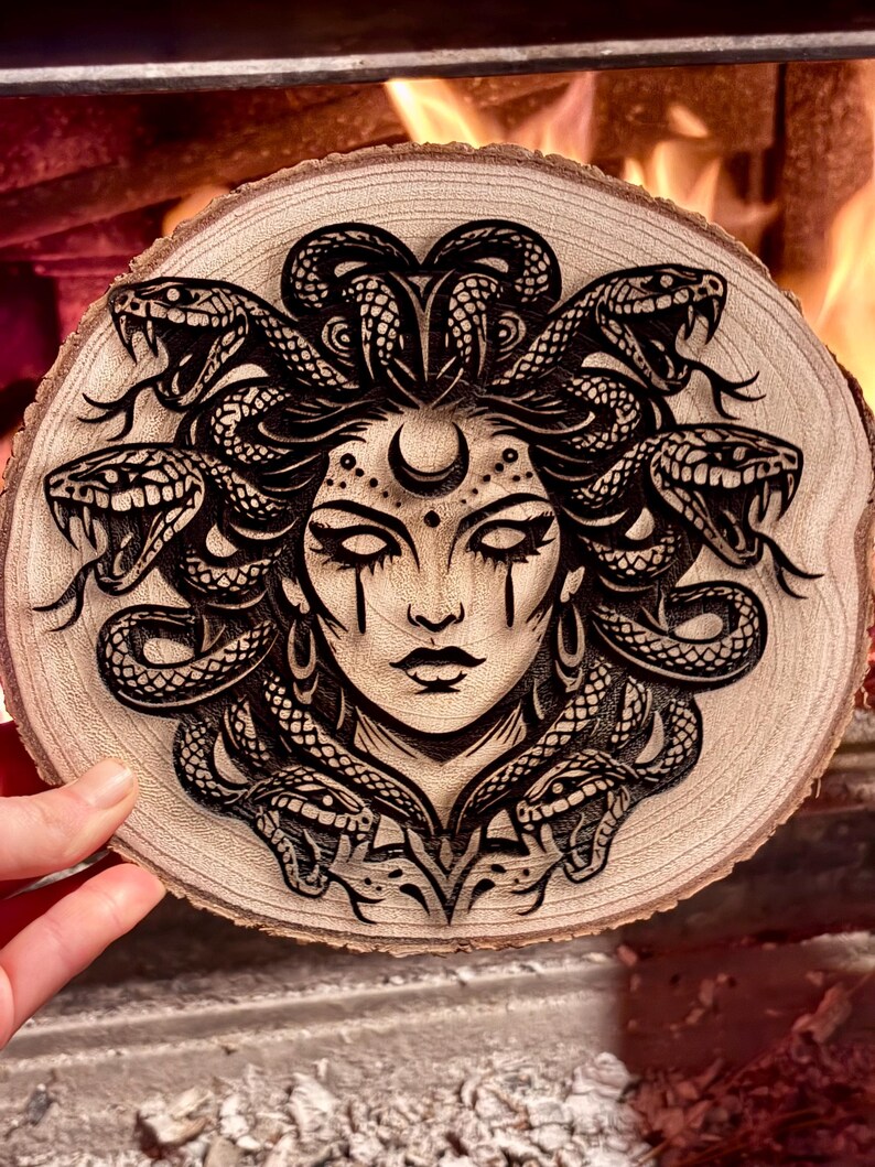 Op de afbeelding: Een houtgebrand kunstwerk met het gezicht van Medusa, met slangen als haar. Het kunstwerk is uitgesneden in een ronde, lichtgekleurde houten schijf. De slangen zijn gedetailleerd en het gezicht heeft een halve maan boven het voorhoofd.