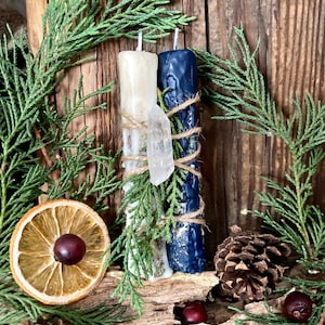 May include: Two cylindrical candles, one cream and one dark blue, are wrapped in twine with a clear quartz crystal and green cedar sprigs. The candles are positioned vertically against a textured wooden surface with dried orange slices and pinecones.