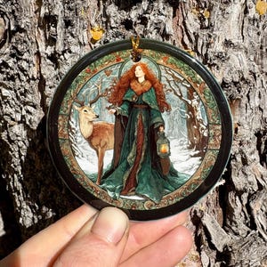 May include: Round ornament featuring a woman with red hair and a green cloak, holding a lantern, standing next to a deer in a snowy forest scene. The ornament has a black border and a gold hanging loop.