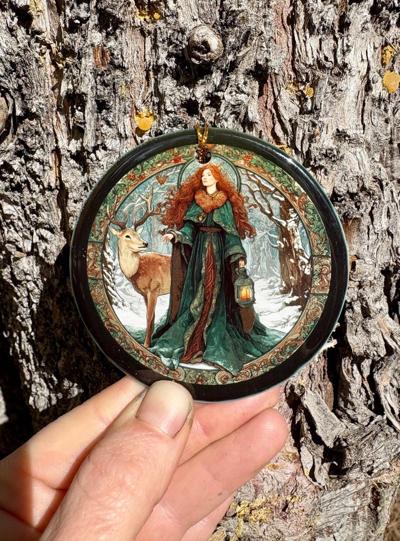 May include: Round ornament featuring a woman with red hair and a green cloak, holding a lantern, standing next to a deer in a snowy forest scene. The ornament has a black border and a gold hanging loop.