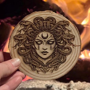 Op de afbeelding: Een ronde houten onderzetter met een gedetailleerd pyrografieontwerp van Medusa met slangen als haar, een maan en ingewikkelde gelaatstrekken. De onderzetter is gemaakt van een natuurlijke houten schijf, met de houtnerf zichtbaar.