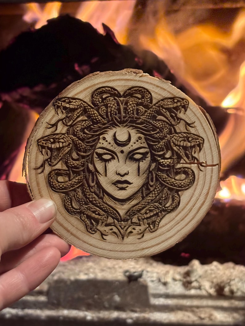 Op de afbeelding: Een ronde houten onderzetter met een gedetailleerd pyrografieontwerp van Medusa met slangen als haar, een maan en ingewikkelde gelaatstrekken. De onderzetter is gemaakt van een natuurlijke houten schijf, met de houtnerf zichtbaar.