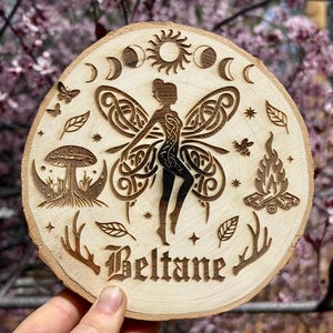 May include: A round wooden plaque with a laser-cut design featuring a fairy with detailed wings, crescent moons, a sun, and the word "Beltane." The plaque also includes a mushroom, leaves, a campfire, and antlers.
