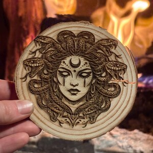 Op de afbeelding: Een ronde houten onderzetter met een gedetailleerde houtgebrande afbeelding van Medusa. Het kunstwerk toont Medusa's gezicht met slangen als haar, een halve maan en traanvormige details. De onderzetter is gemaakt van lichtgekleurd hout.