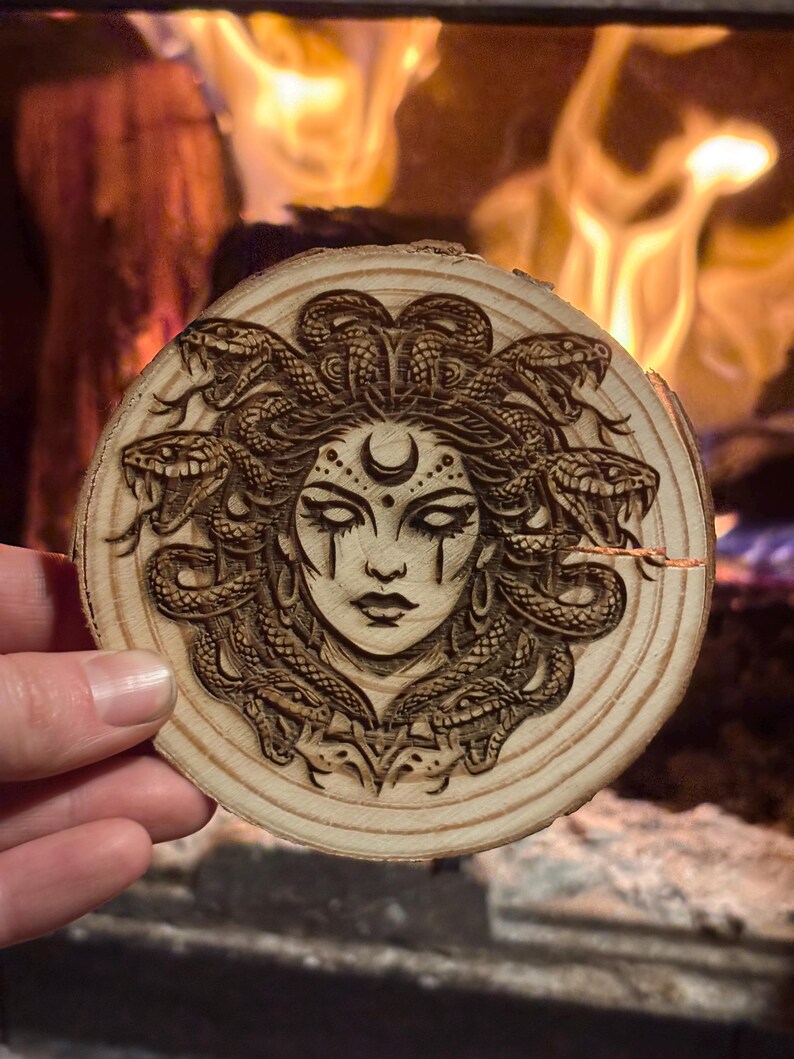 Op de afbeelding: Een ronde houten onderzetter met een gedetailleerde houtgebrande afbeelding van Medusa. Het kunstwerk toont Medusa's gezicht met slangen als haar, een halve maan en traanvormige details. De onderzetter is gemaakt van lichtgekleurd hout.