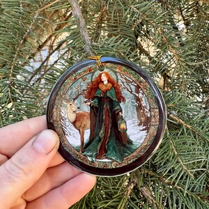 May include: A round Christmas ornament featuring a woman with red hair and a deer in a winter forest scene. The woman is wearing a green and red dress and holding a lantern. The ornament has a gold string for hanging.