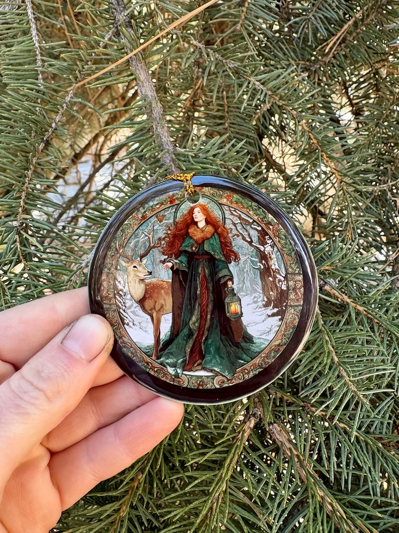 May include: A round Christmas ornament featuring a woman with red hair and a deer in a winter forest scene. The woman is wearing a green and red dress and holding a lantern. The ornament has a gold string for hanging.