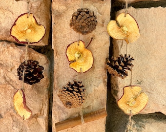 May include: A rustic wall hanging featuring dried apple slices, pine cones, a cinnamon stick, and small brass bells suspended from a wooden branch. The natural elements are attached with twine, creating a charming, earthy aesthetic.
