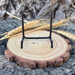 May include: A black metal display stand featuring two vertical uprights connected by a horizontal base bar, designed with two small front-facing hooks at the bottom to support items in an upright position for viewing or decorative display purposes.