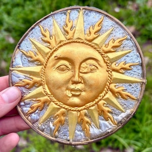 May include: A round, golden sun-faced decorative piece with a detailed design. The sun has a smiling face, surrounded by golden rays and flames, set against a silver and blue background. The piece is held by a hand.