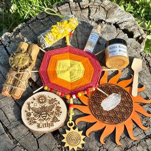 May include: An assortment of spiritual and ritual items, including a colorful yarn mandala, a wooden sunburst, a jar of scrub, a wooden disc with a bee design, and a bundle of dried herbs. The items are arranged on a tree stump.