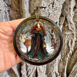 May include: Round ceramic ornament featuring a woman with red hair in a green gown, holding a lantern, and standing next to a deer in a snowy forest scene. The ornament has a gold hanging loop.