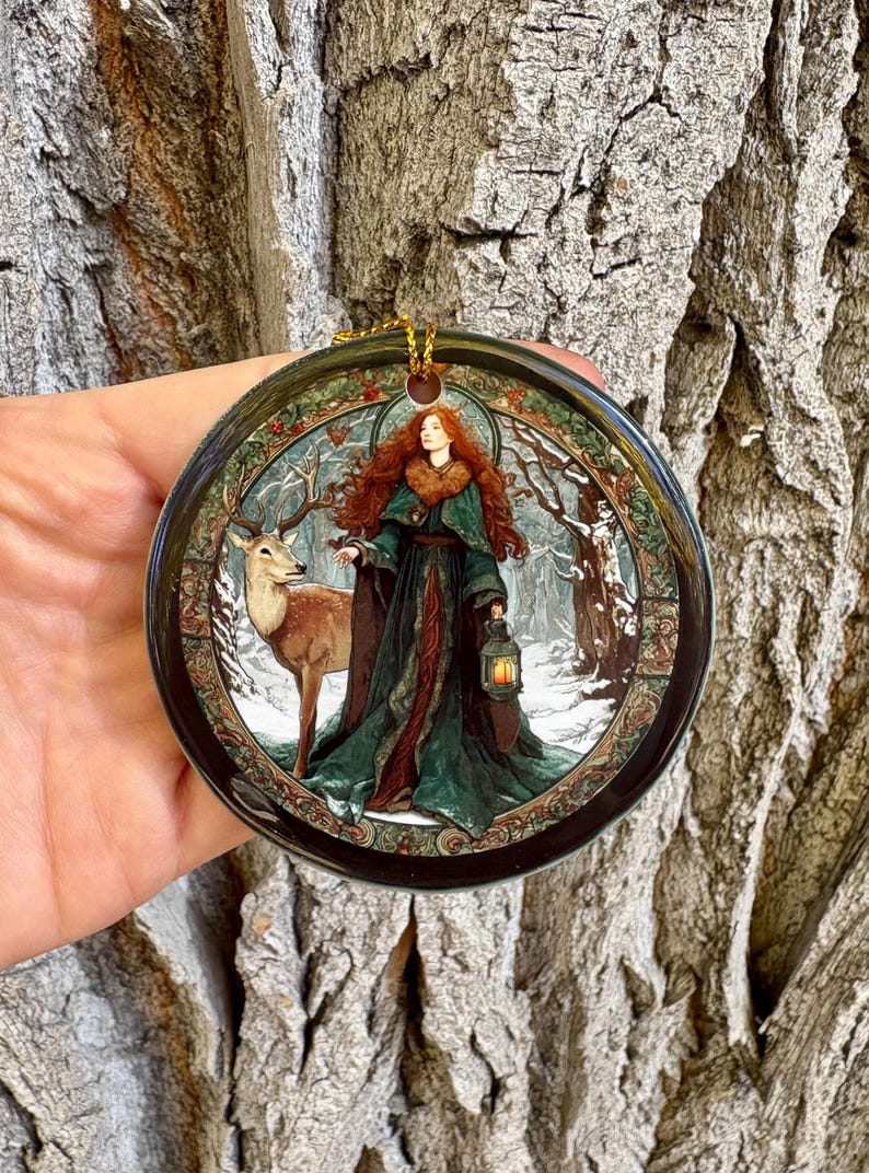 May include: Round ceramic ornament featuring a woman with red hair in a green gown, holding a lantern, and standing next to a deer in a snowy forest scene. The ornament has a gold hanging loop.
