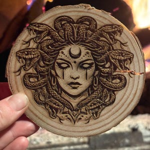 Op de afbeelding: Een ronde houten onderzetter met een gedetailleerd laserge&euml;tst ontwerp van Medusa's gezicht. Het kunstwerk toont slangen als haar, een halve maan en traandruppel details. De onderzetter is gemaakt van een natuurlijke houten schijf.