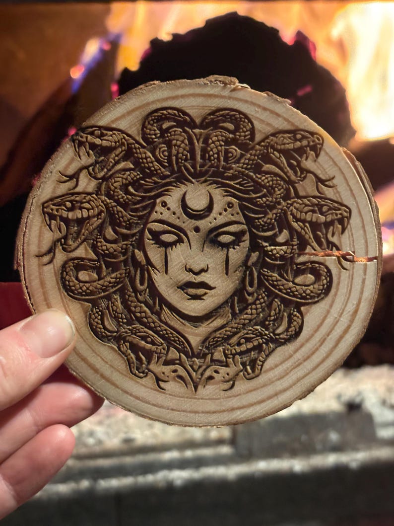 Op de afbeelding: Een ronde houten onderzetter met een gedetailleerd laserge&euml;tst ontwerp van Medusa's gezicht. Het kunstwerk toont slangen als haar, een halve maan en traandruppel details. De onderzetter is gemaakt van een natuurlijke houten schijf.