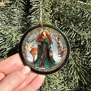 May include: A round Christmas ornament featuring a woman with red hair and a green dress, holding a lantern, standing next to a deer in a snowy forest scene. The ornament has a black border and a gold string for hanging. The background is a green Christmas tree.