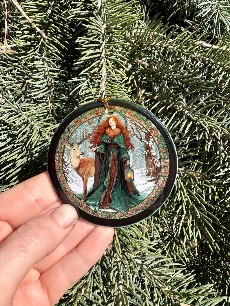 May include: A round Christmas ornament featuring a woman with red hair and a green dress, holding a lantern, standing next to a deer in a snowy forest scene. The ornament has a black border and a gold string for hanging. The background is a green Christmas tree.