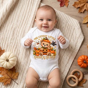 May include: A white long-sleeve baby bodysuit featuring a graphic of a teddy bear wearing a pumpkin hat, sitting among pumpkins, a ghost, and a bat. The text reads THE CUTEST LITTLE PUMPKIN IN THE PATCH in orange and brown lettering.