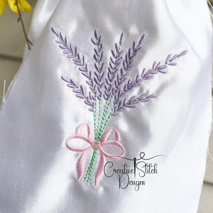Könnte beinhalten: Ein weißer Stoff mit einer gestickten Lavendel-Bouquet mit einer rosa Schleife. Der Text "Creative Stitch Designs" ist unter dem Bouquet gestickt.