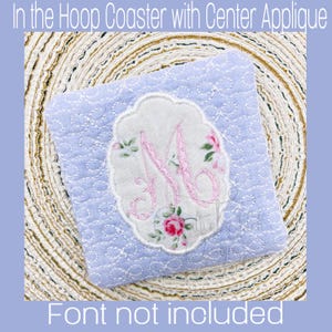 May include: A light blue fabric coaster with a white oval applique featuring a pink embroidered letter "M" and floral details. The coaster is on a brown woven surface. "Font not included" is written below the coaster.