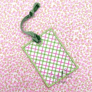 May include: A rectangular gift tag with a green and pink plaid pattern, outlined in green stitching. A green velvet ribbon is attached at the top. The tag has the text "Creative Stitch Designs" in a script font.