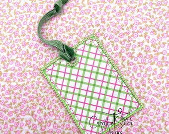 May include: A rectangular gift tag with a green and pink plaid pattern, outlined in green stitching. A green velvet ribbon is attached at the top. The tag has the text "Creative Stitch Designs" in a script font.
