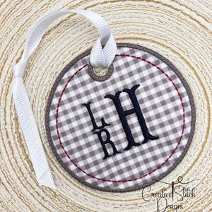 May include: A round, embroidered ornament with a white and gray gingham pattern. The ornament features the embroidered initials "LHR" in black, a brown border, and a red stitched accent. A white ribbon is attached for hanging. The words "Creative Stitch Designs" are at the bottom.