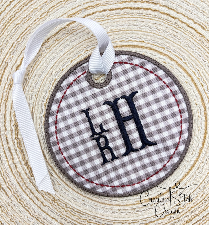 May include: A round, embroidered ornament with a white and gray gingham pattern. The ornament features the embroidered initials "LHR" in black, a brown border, and a red stitched accent. A white ribbon is attached for hanging. The words "Creative Stitch Designs" are at the bottom.