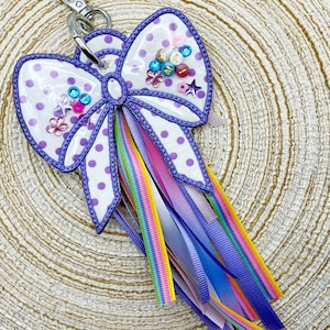 May include: A white and purple polka dot bow-shaped keychain with a silver clasp. The bow has a purple outline and is decorated with colorful gems and ribbons in shades of purple, pink, and rainbow colors. The keychain has the letters "A", "L", and "M" on it.
