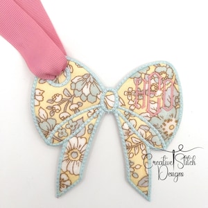 May include: A yellow and blue floral fabric bow with a pink monogram "HCG" embroidered on it. The bow is attached to a pink ribbon.