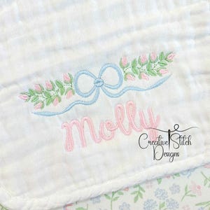 May include: White baby blanket with embroidered details. Features a light blue bow and floral design with the name "Molly" in pink script. The blanket has a floral pattern on the bottom right corner and the text "Creative Stitch Designs".