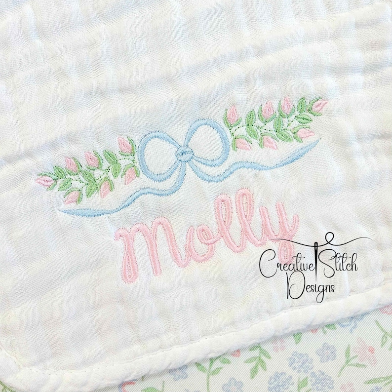 May include: White baby blanket with embroidered details. Features a light blue bow and floral design with the name "Molly" in pink script. The blanket has a floral pattern on the bottom right corner and the text "Creative Stitch Designs".