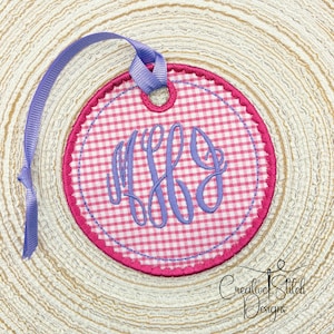 Könnte beinhalten: Ein Kreis aus rosa-weißem Gingham-Stoff mit einem gewellten Rand und einem violetten Monogramm in der Mitte. Der Kreis ist an einem violetten Band befestigt.
