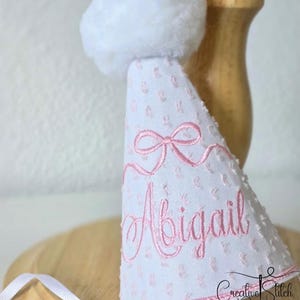May include: A white cone-shaped party hat featuring a textured swiss dot fabric with pink accents. The hat is embroidered with a pink bow and the name Abigail in cursive script. A large white pom-pom sits on top, and a thin white ribbon is attached.
