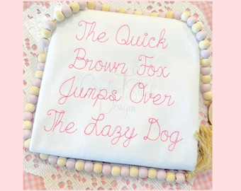 May include: A white fabric with pink embroidery of the phrase "The Quick Brown Fox Jumps Over The Lazy Dog" in a cursive font. The fabric is surrounded by a string of light brown and purple beads.
