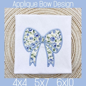 May include: A blue and white floral applique bow design for embroidery. The bow is outlined in blue and has a floral pattern with blue flowers on a white background.