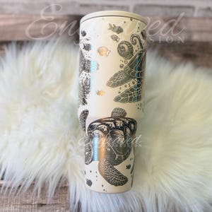A white ceramic mug with a black and silver turtle design printed on it, placed on a white furry surface.