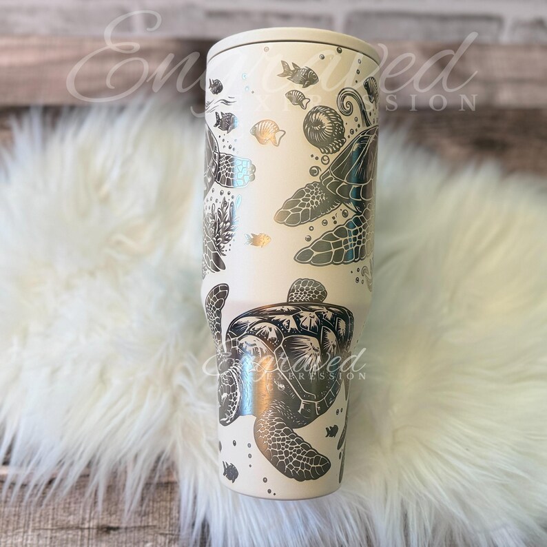 A white ceramic mug with a black and silver turtle design printed on it, placed on a white furry surface.
